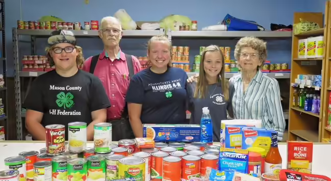 three teen 4-H members and two adults standing in food pantry with collection of food on counter